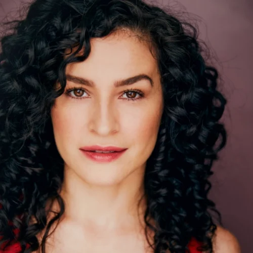 Woman with long, dark curly hair and light skin looks directly at the camera with a neutral expression, against a plain, muted background.