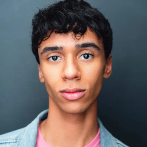 A young person with short, dark hair poses against a plain, dark background, wearing a light denim jacket over a pink shirt.