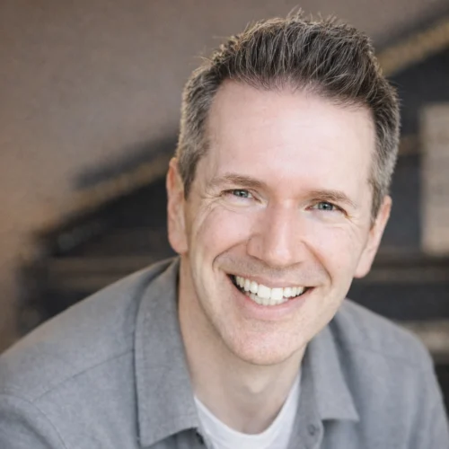 Dominique Kelley, a man with short brown hair, wearing a gray shirt over a white t-shirt, smiles at the camera in an indoor setting.