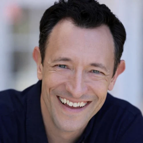 A man with short dark hair and blue eyes is smiling at the camera, wearing a dark shirt, with a blurred light background.