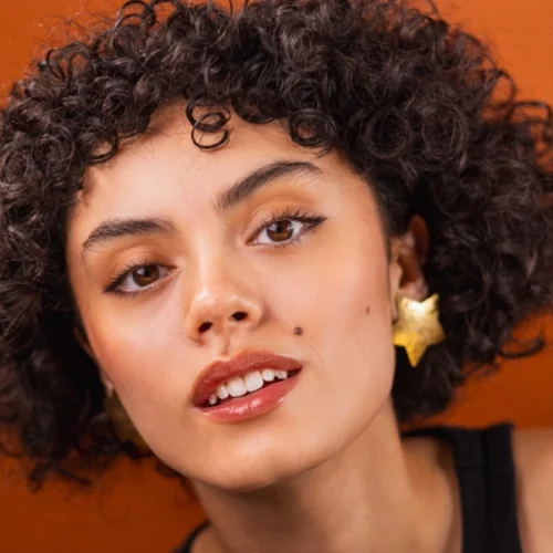 A person with short curly hair, wearing star-shaped earrings and a black top, poses in front of an orange background.