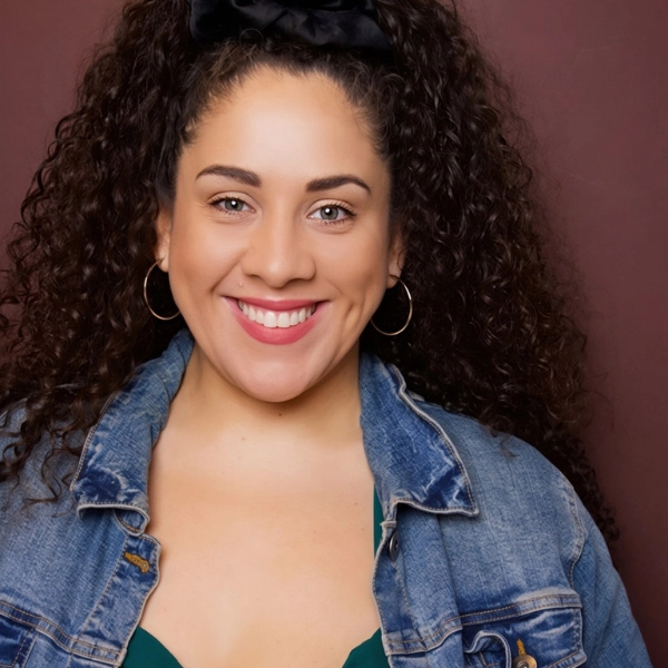 A woman with curly hair, wearing hoop earrings, a denim jacket, and a green top, smiles at the camera against a maroon background.