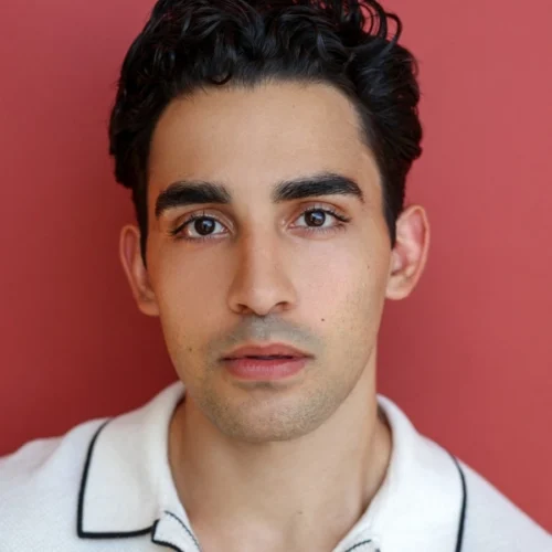 A young man with short, dark curly hair and brown eyes faces the camera against a plain red background, wearing a white collared shirt with black trim.