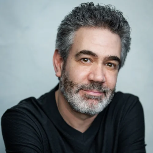 A middle-aged man with gray hair and a beard, wearing a black shirt, sits against a plain light background and looks at the camera with a slight smile.