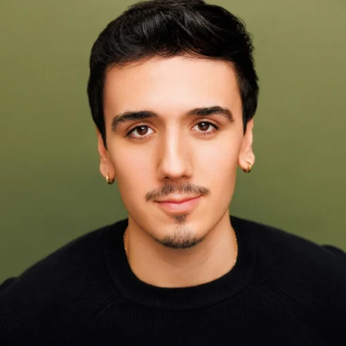 A young man with short dark hair, a trimmed mustache and goatee, and gold hoop earrings, wearing a black sweater, poses against a plain olive green background.