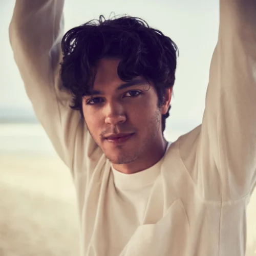 A young man with wavy dark hair and light facial hair looks at the camera, wearing a light-colored shirt, with his arms raised against a blurred outdoor background.