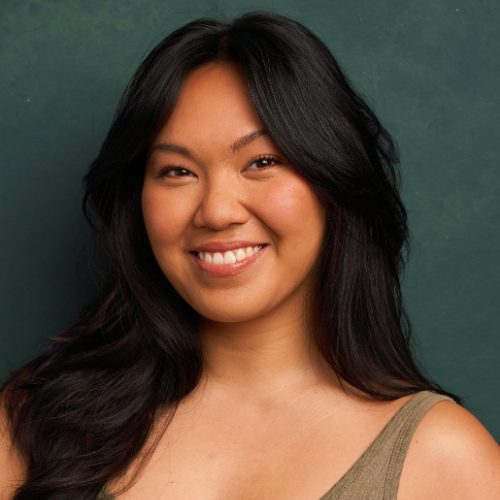 A woman with long black hair and a sleeveless top smiles in front of a green background.