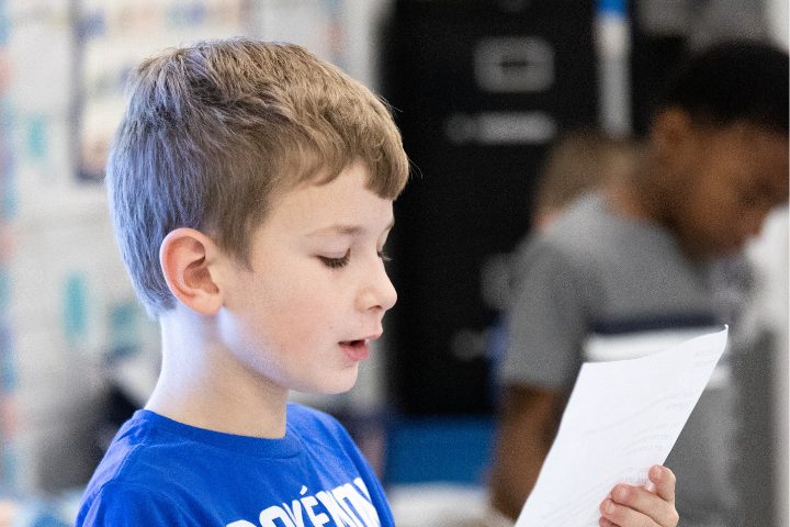 A young boy in a blue shirt reads from a sheet of paper in a classroom setting, with another student blurred in the background.