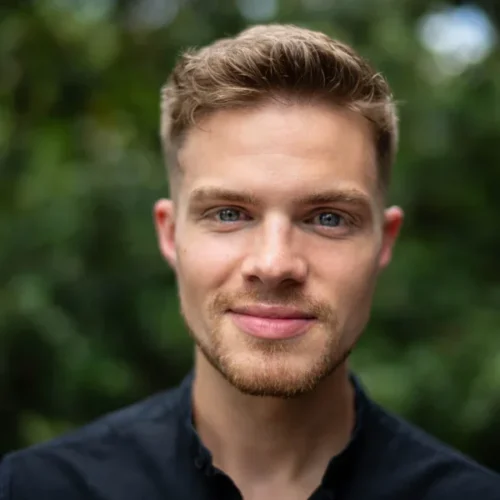 A young man with short blond hair, a trimmed beard, and blue eyes, wearing a black button-up shirt, stands outdoors with greenery in the background.