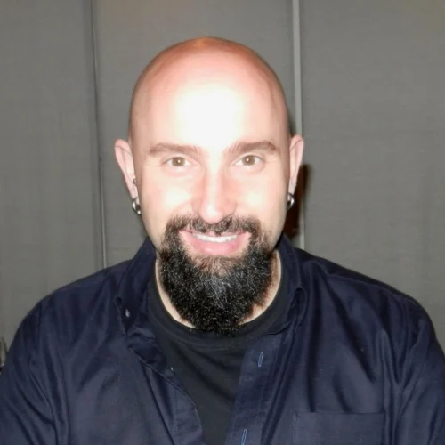 A bald man with a black goatee and mustache, wearing a dark shirt, smiles at the camera against a plain background.