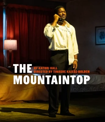 A man stands on stage in front of a bed, holding a phone to his ear. Text on image reads: "The Mountaintop by Katori Hall, directed by Tinashe Kajese-Bolden." Experience this powerful moment with Dinosaur Stream.