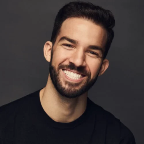A man with short dark hair and a beard smiles broadly, showing his teeth. He is wearing a black shirt and is posed against a dark background.