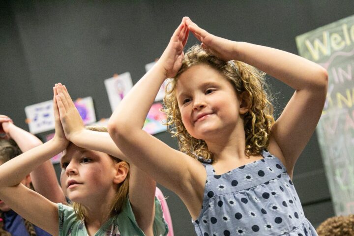 Two young girls in a classroom stand with their hands forming a triangle above their heads. A chalkboard with writing and children's artwork are visible in the background.