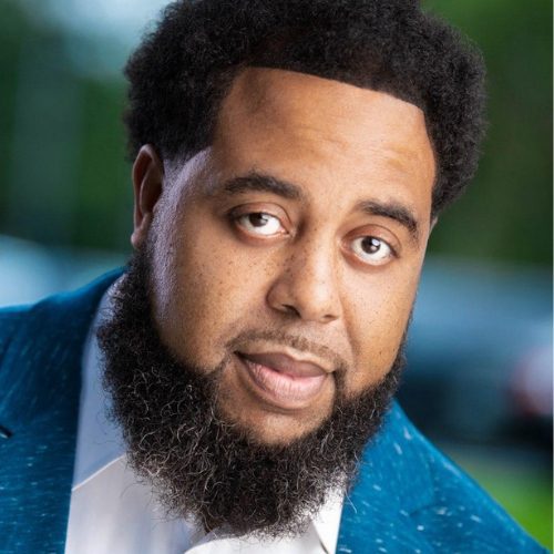 A man with a full beard and short, curly hair wears a blue suit jacket and white shirt, looking directly at the camera with a slight smile.