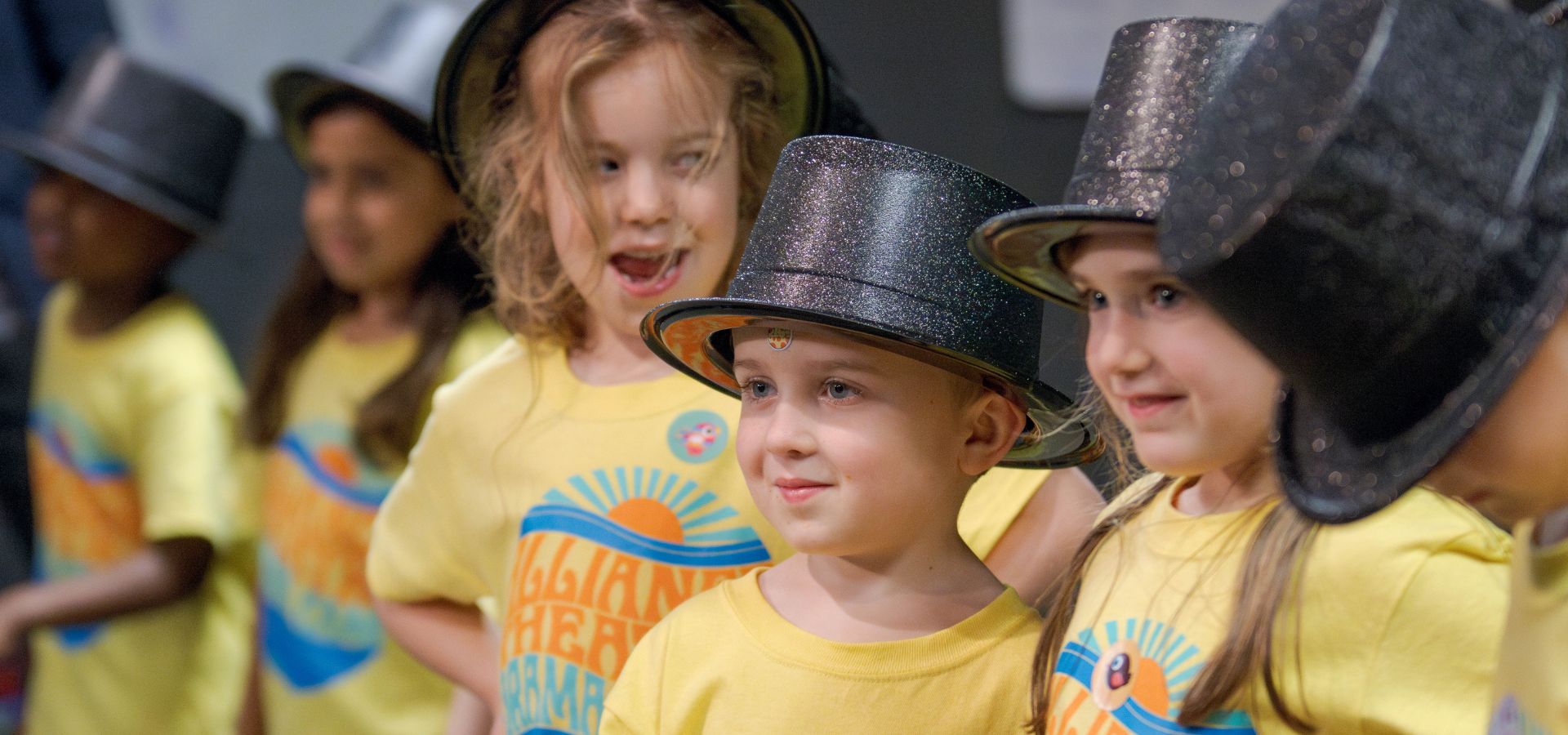 A group of young children wearing yellow t-shirts and black top hats stand together, some smiling, during what appears to be a drama camp performance or event.