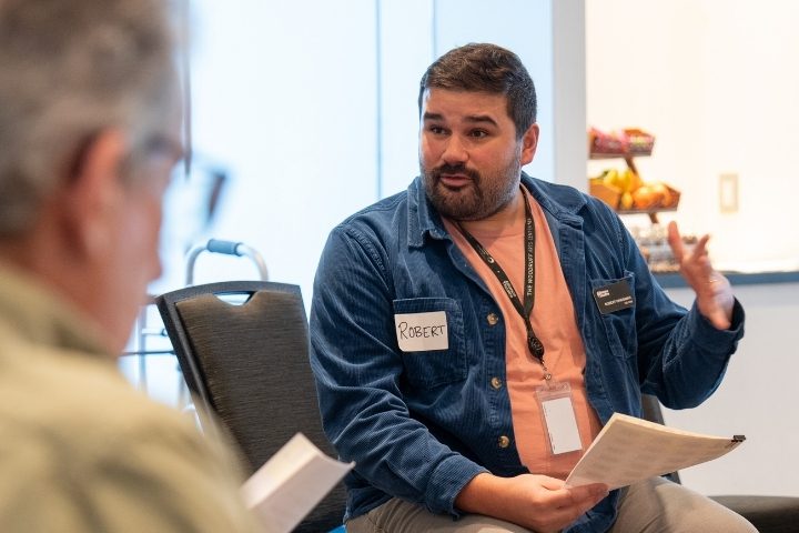 A man wearing a name tag labeled "Robert" speaks while holding papers in a meeting room; another person is partially visible in the foreground.