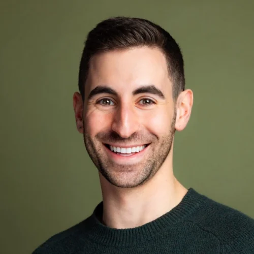 A man with short dark hair and a trimmed beard, wearing a green sweater, smiles in front of an olive green background.