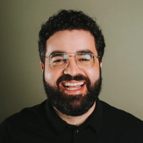 A man with curly dark hair, a beard, and glasses is smiling broadly at the camera. He is wearing a black shirt and is posed in front of a plain, light-colored background.