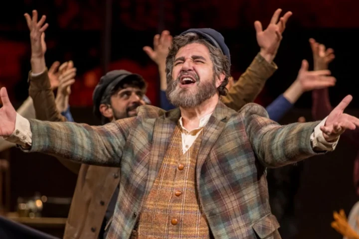 A man in plaid clothing stands center stage with arms outstretched, surrounded by other performers with raised hands during a theatrical performance, reminiscent of a moment from Fiddler on the Roof: A Behind-the-Scenes Feature.