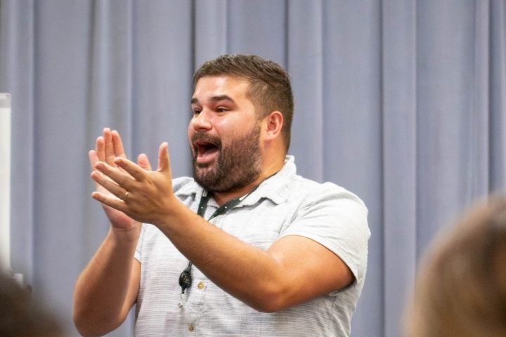A bearded man in a short-sleeve shirt enthusiastically claps his hands while speaking in front of a gray curtain backdrop.