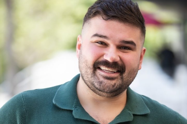 Man with short dark hair and beard, wearing a green collared shirt, smiles outdoors with a blurred background.