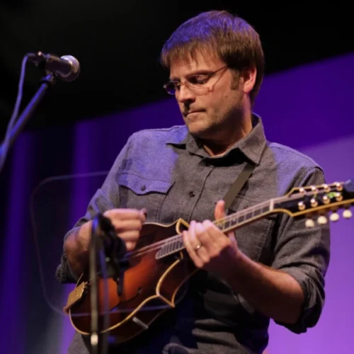 A man wearing glasses and a button-up shirt plays a mandolin on stage in front of a microphone.