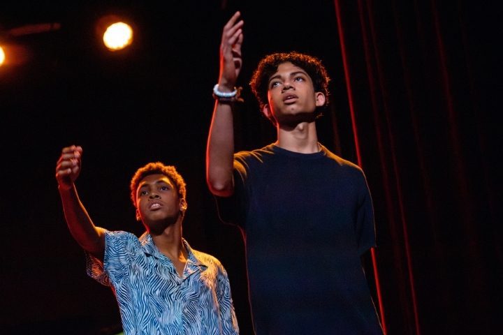Two young men stand on stage under spotlights, each with one arm raised and gazing upward, performing against a dark background.