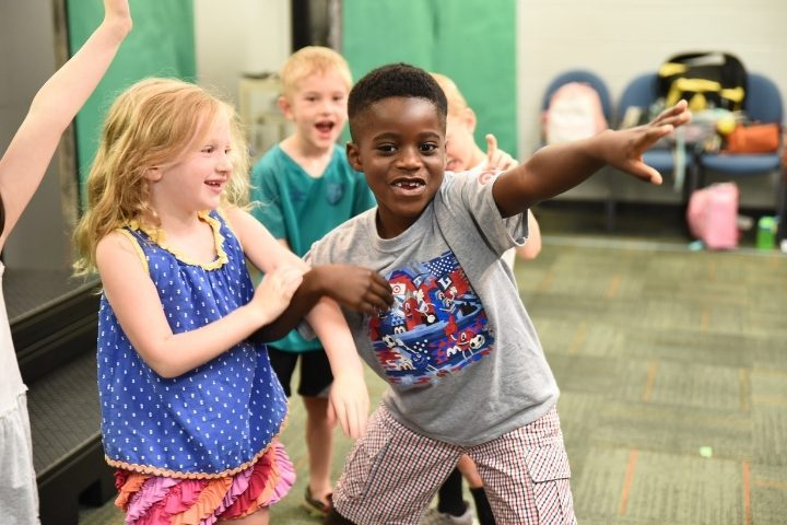 A group of children stand together indoors at the Alliance Theatre, smiling. One boy in front extends his arm outward while others laugh and interact around him.