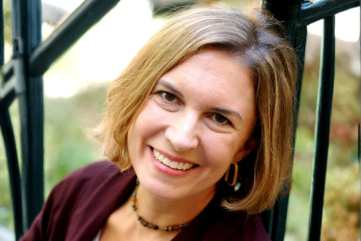 Announcing the Creative Team of Basura: A woman with short blonde hair smiles at the camera, wearing a maroon jacket, beaded necklace, and gold hoop earrings, standing near a metal railing outdoors.