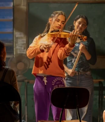 A young woman in an orange sweatshirt and purple sweatpants plays the violin in a classroom during an A Christmas Carol Stream, with two other people seated nearby and one person standing in the background.