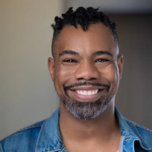 Smiling man with short curly hair and a beard, wearing a denim jacket, stands indoors against a neutral background—this is likely Dwight Andrews.