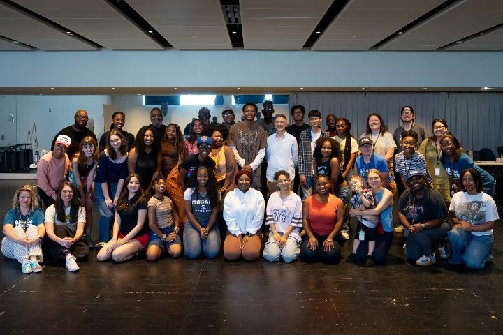 A large group of people poses together in a rehearsal room, smiling at the camera.