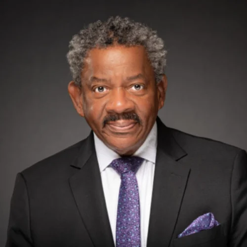 An older man with gray hair wearing a black suit, white shirt, and purple patterned tie poses against a dark background. This distinguished portrait captures Dwight Andrews in a moment of quiet confidence and elegance.