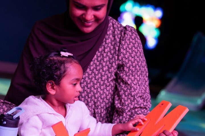 A woman smiles as a young girl in a white hoodie examines large orange foam letters.