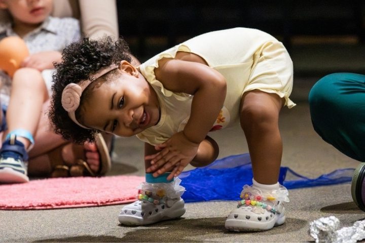 A toddler in a yellow outfit and headband bends down and smiles, surrounded by other children and adults.