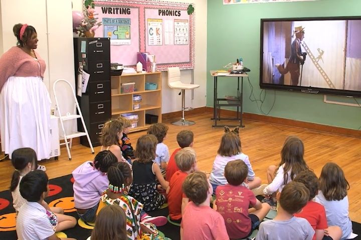 A group of young children sits on the floor of a classroom watching a musical on a large screen, with a teacher standing nearby. Writing and phonics posters are displayed on the wall.