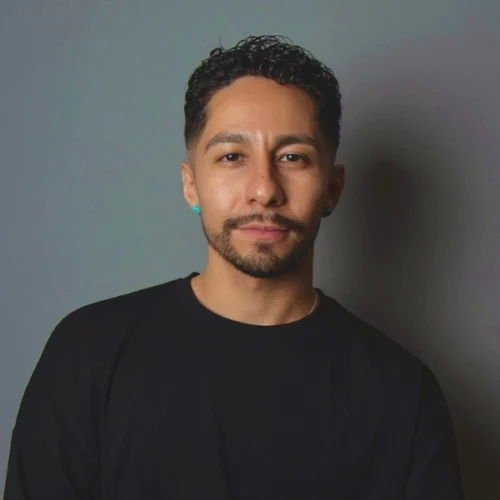 Man with short curly hair, trimmed beard, and mustache, wearing a black shirt and turquoise earrings, stands against a plain gray background, looking directly at the camera with a neutral expression.