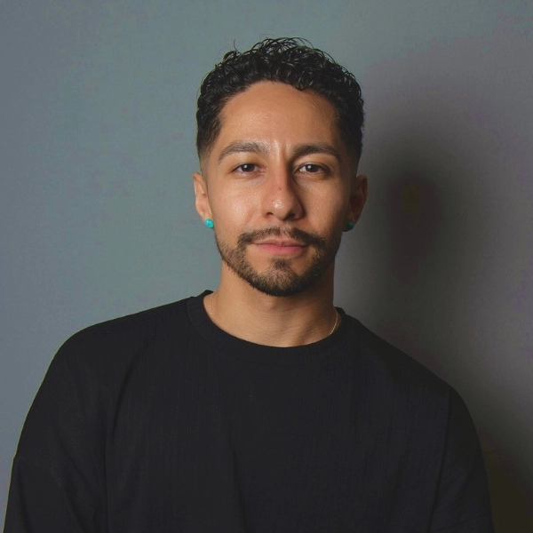 Man with short curly hair, trimmed beard, and mustache, wearing a black shirt and turquoise earrings, stands against a plain gray background, looking directly at the camera with a neutral expression.