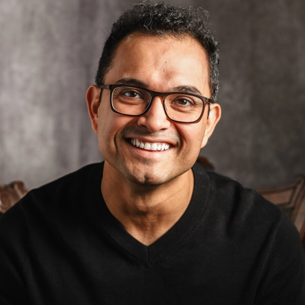 A person with short black hair and glasses smiles while wearing a black sweater, seated in front of a neutral gray background.