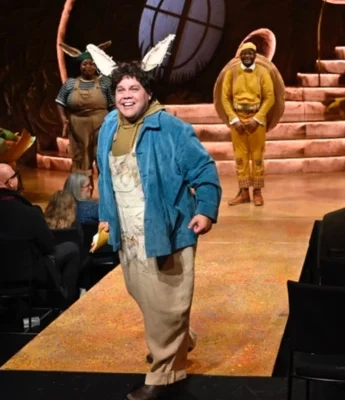 An actor in donkey ears and costume walks on stage, with two other actors in animal costumes and an abstract set in the background—capturing a playful theatrical spirit reminiscent of an A Christmas Carol Stream. Audience members are seated nearby.