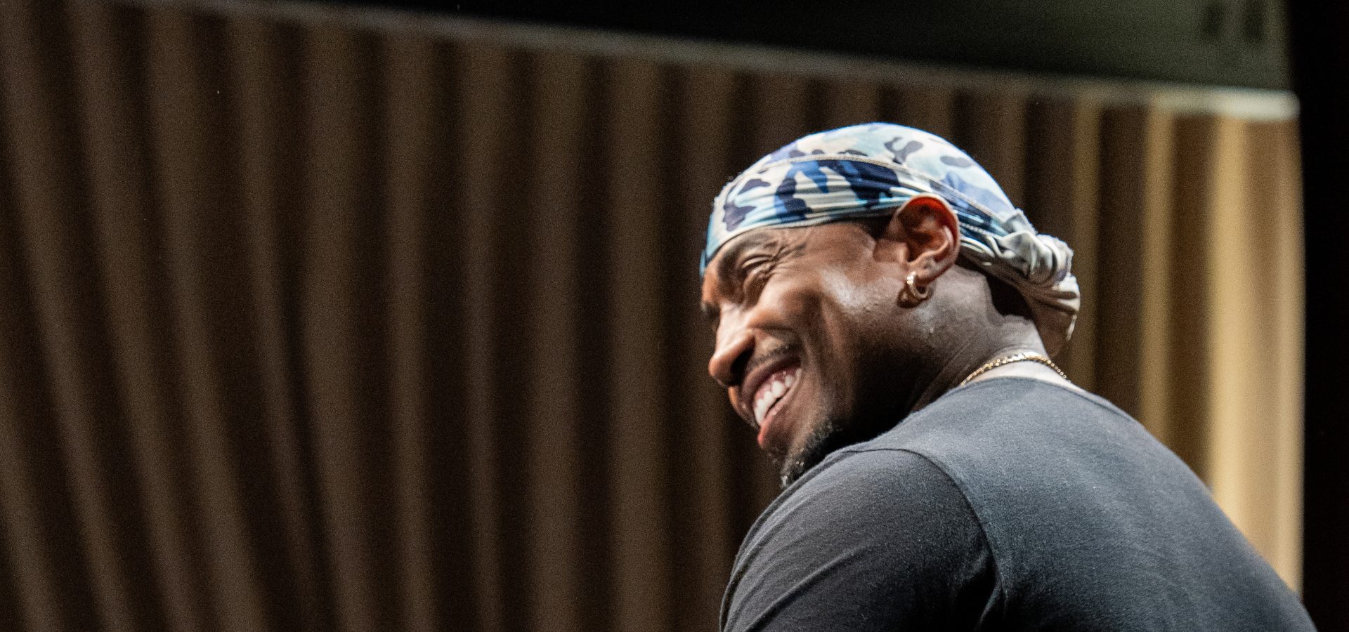 A man wearing a blue and white bandana and a black shirt smiles while looking to the side with a background of curtain panels.