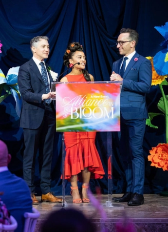 Three people stand on stage behind a colorful “Alliance in Bloom” podium, dressed formally, with large paper flowers in the background.
