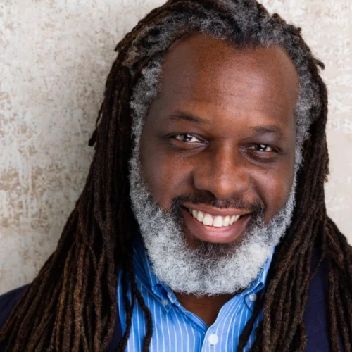 Smiling man with long dark dreadlocks and a full gray beard, wearing a blue striped shirt and dark jacket, posed against a textured beige background in a portrait by Amanda Washington.