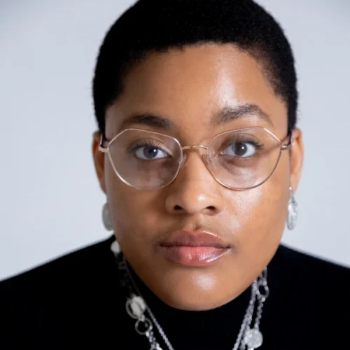 Amanda Washington, a person with short hair, glasses, and earrings, looks directly at the camera, wearing a black top and layered necklaces against a plain background.