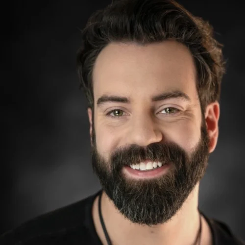 A man with dark hair and a full beard smiles at the camera against a dark background, reminiscent of the professional portraits often taken by Rachel Sabo-Hedges.