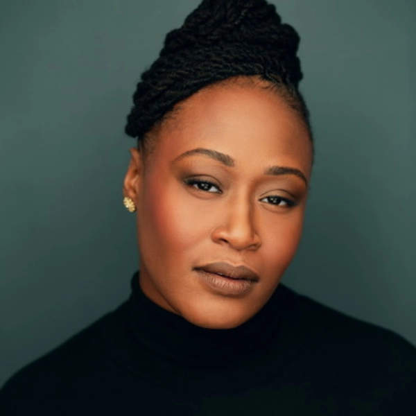A woman with braided hair styled in an updo, wearing a black turtleneck and stud earrings, poses against a plain dark background.