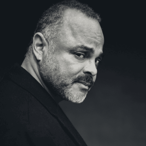 A man with short hair and a beard looks over his shoulder, wearing a dark jacket against a dark background.