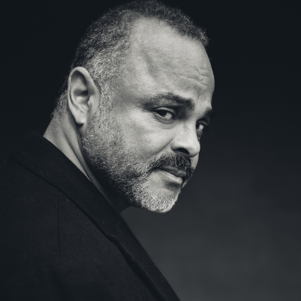 A man with short hair and a beard looks over his shoulder, wearing a dark jacket against a dark background.