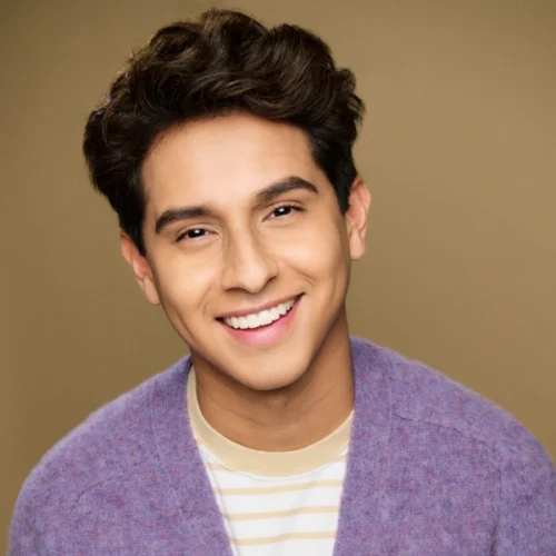 Young person with short, wavy dark hair smiles at the camera, wearing a light purple sweater over a beige and white striped shirt, against a plain tan background.