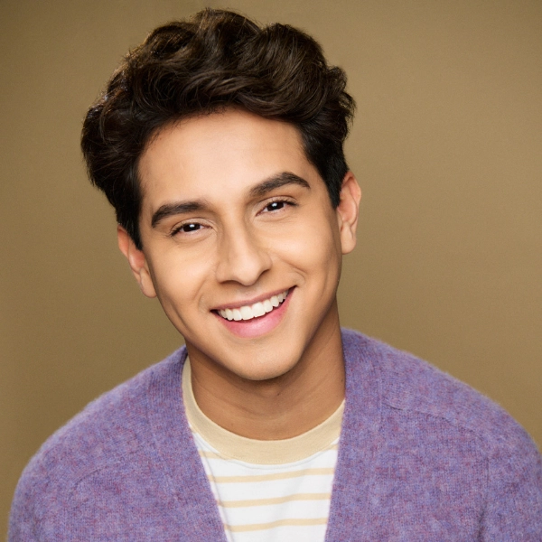 Young person with short, wavy dark hair smiles at the camera, wearing a light purple sweater over a beige and white striped shirt, against a plain tan background.